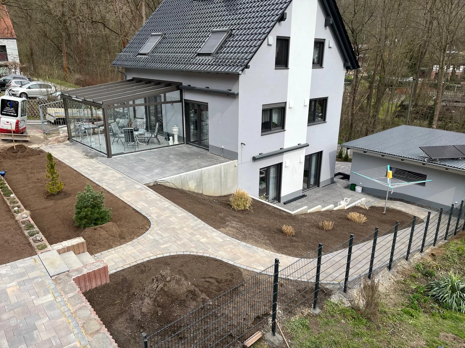 Neu angelegte Gartenanlage mit geschwungenem Pflasterweg, frischen Beeten, Rasenflächen und Doppelstabmattenzaun an einem Einfamilienhaus mit Wintergarten